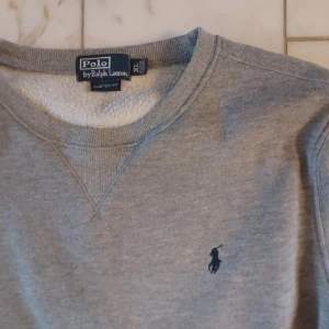 Grå tröja från Polo Ralph Lauren - Säljer en klassisk grå tröja från Polo Ralph Lauren. Den har en broderad logga på bröstet och är i mycket bra skick. Perfekt för både höst och vår, med långa ärmar och en bekväm passform. Tröjan är gjord i mjukt material som håller dig varm och stilren. Inga tecken på användning. XL men passar bäst M eller L