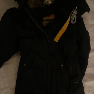 Svart vinterjacka med pälsdetaljer - Säljer en svart Parajumpers vinterjacka i mycket bra skick. Jackan har en mysig pälsfodrad huva och praktiska fickor. Den är perfekt för kalla vinterdagar och har en snygg patch på ärmen. Dragkedja och knappar framtill för extra skydd mot kylan.