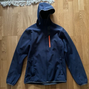 Mörkblå jacka från McKinley - Säljer en mörkblå jacka från McKinley i bra skick. Den har en orange dragkedja på bröstet och en praktisk huva. Perfekt för höst och vår, med långärmade ärmar och en stilren design. Passar bra för utomhusaktiviteter och vardagsbruk.