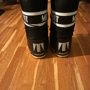 Svarta Moon Boots - Säljer ett par svarta Moon Boots i mycket bra skick. De är perfekta för vintern med sin varma och bekväma design. Skorna har snörning och en vit logga runt skaftet. De är gjorda i syntetmaterial och har en robust sula för bra grepp på snöiga underlag. 
