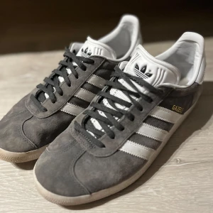 Adidas Gazelle 41  - Säljer ett par snygga grå Adidas Gazelle sneakers i mocka. Storlek 41 & 1/3. De har de klassiska vita ränderna och grå / svart snörning. Perfekta för både vardag och fest! Skorna är i mycket bra skick och redo för nya äventyr.