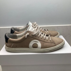 Oliver Cabbel low fog 1 - Säljer ett par snygga beige sneakers med vit cirkeldetalj på sidan. Skorna har snörning och en vit sula. De är i bra skick med lite slitage på sulan. Perfekta för vardagsbruk och passar till många olika outfits. Ord pris inkl tull 4000kr mitt pris endast 1000!!! 