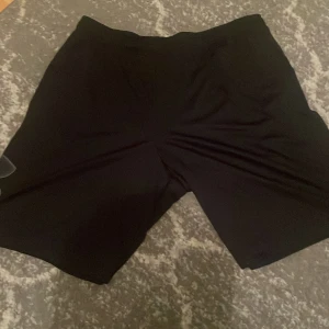 Svarta shorts från Under Armour - Säljer ett par svarta shorts från Under Armour i storlek XL. Det är helt nya och aldrig använda och har en loose passform, perfekt för träning. Materialet är lätt och andas bra, vilket gör dem idealiska för sommarens träningspass. Stort logotyptryck på benet ger en sportig look.