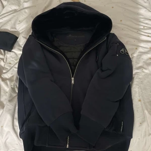 Mooske Knuckles Classic Bunny - Säljer en snygg mörkblå classic bunny från Moose knuckles i mycket bra skick. Den har en cool design med dragkedja och en praktisk huva. Jackan är quiltad på insidan för extra värme och har en unik detalj på ärmen. Perfekt för höst och vinter!