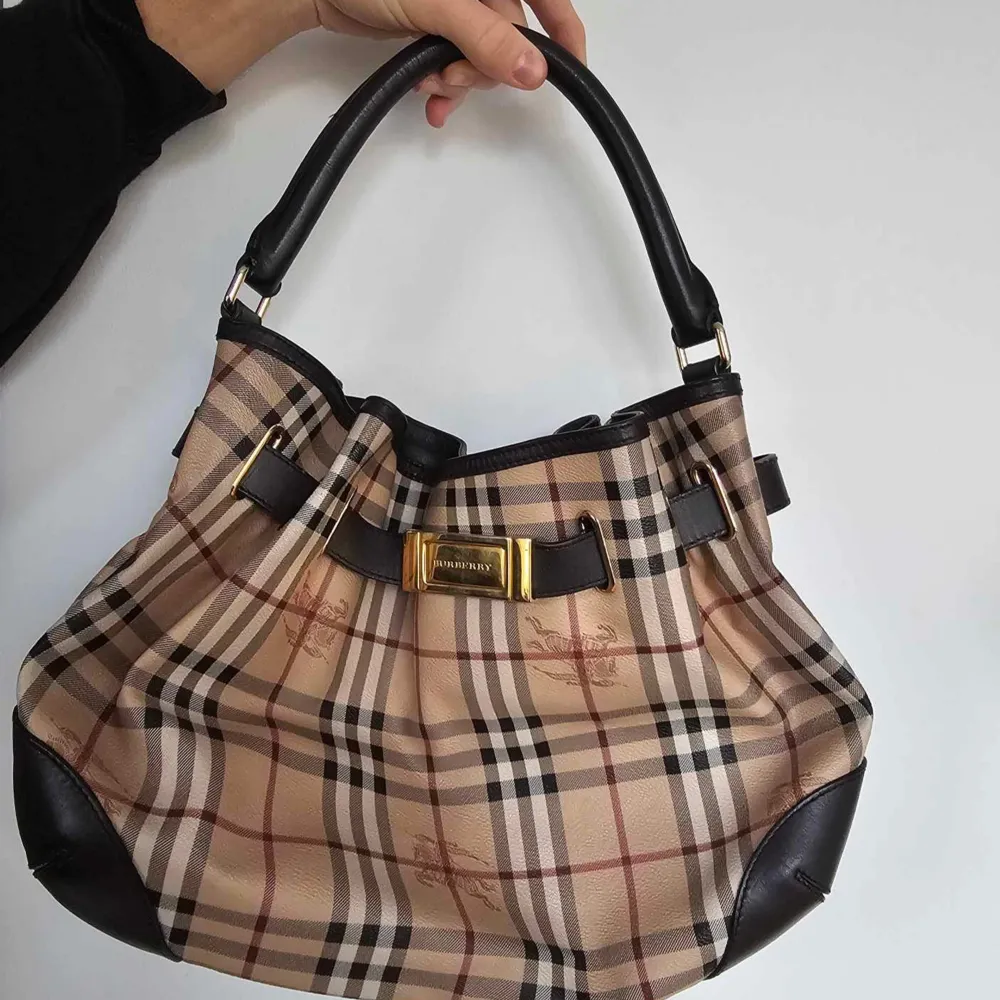 Säljer en snygg  burberry rutig handväska med klassiskt mönster och bruna läderdetaljer. Väskan har en elegant rosett och guldfärgad spänne framtill. Perfekt för både vardag och fest!. Väskor.