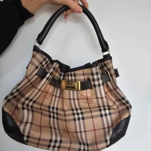 Äkta burberry handväska - Säljer en snygg  burberry rutig handväska med klassiskt mönster och bruna läderdetaljer. Väskan har en elegant rosett och guldfärgad spänne framtill. Perfekt för både vardag och fest!
