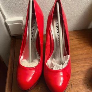 Röda pumps från Rainbow - Snygga röda pumps från Rainbow i storlek 41.Har tyvärr ett svart märke och vet ej om det går att få bort. De har en hög klack och är i tillfredsställande skick. Perfekta för festliga tillfällen när du vill sticka ut lite extra. Skorna har en glansig finish som ger en lyxig känsla.