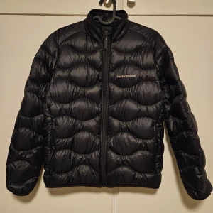 Svart pufferjacka från Peak Performance - Säljer en snygg svart pufferjacka från Peak Performance i mycket bra skick. Jackan är perfekt för kalla dagar med sitt quiltade mönster och dragkedja framtill. Den har långa ärmar och är gjord i ett slitstarkt syntetmaterial. Perfekt för både höst och vinter!