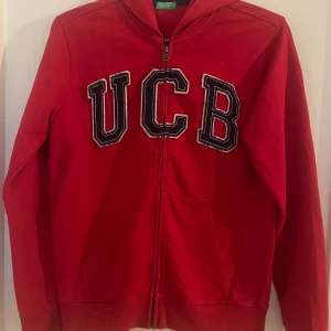 Röd hoodie med UCB-logga - Säljer en snygg röd hoodie från United Colors of Benetton med stor UCB-logga på framsidan. Den har dragkedja och är perfekt för både vardag och chill. Superbekväm och i bra skick!