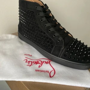 Svarta sneakers från Christian Louboutin - Säljer ett par svarta sneakers från Christian Louboutin i mycket bra skick. Använda en gång på balen. HELT ÄKTA. De är täckta med svarta nitar och har en cool, edgy look. Skorna har snörning och är perfekta för en fest eller en utekväll. De är gjorda av mocka och har en klassisk röd sula som är typisk för märket. Mer bilder skriv meddelande att önskar mer bilder. 
