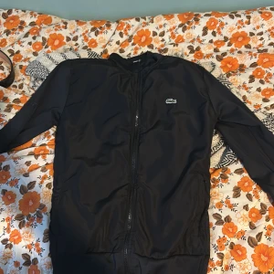 Svart jacka från Lacoste - Säljer en snygg svart jacka från Lacoste i storlek M. Jackan har en dragkedja framtill och det klassiska Lacoste-logotypen på bröstet. Perfekt för höst och vår, med en stilren design som passar till det mesta. Skön och enkel att bära, idealisk för vardagsbruk.