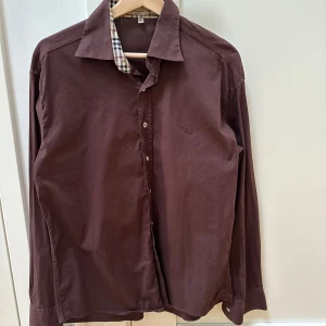Plommonlila/brun Burberry skjorta storlek L - Säljer en snygg skjorta från Burberry i slim fit, storlek L. Skjortan har långa ärmar och klassiska Burberry-rutor på insidan av kragen. Perfekt för både vardag och fest! Skjortan är i nyansen ngt plommonfärgad och drar ngt även åt chokladbruna hållet beroende på ljus.