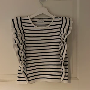 Randig topp från ONLY - Säljer en superfin randig topp från ONLY i storlek M. Den är vit med svarta ränder och har volangdetaljer längs sidorna. Perfekt för både vardag och fest! 🖤🤍