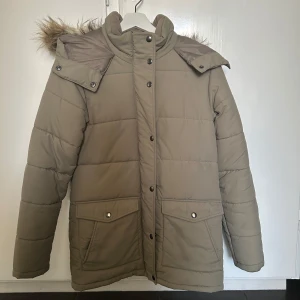 Beige pufferjacka med fuskpäls - Säljer en snygg och varm beige pufferjacka i jättefint skick. Jackan har långa ärmar och praktiska fickor med knappar. Perfekt för kalla vinterdagar! Enda skavanken är att ögglan man hänger upp jackan i har gått av på ena sidan, men den går enkelt att sy dit igen om man vill hänga jackan på krok istället för på galge.