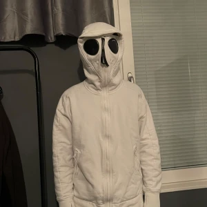 C.P. Company hoodie - Säljer en cool vit jacka från C.P. Company i storlek M. Jackan har en unik design med huva och inbyggda glasögon, perfekt för en futuristisk look. Den är i mycket bra skick och har dragkedja framtill samt praktiska fickor med dragkedjor. Perfekt för höst och vår!
