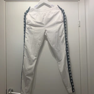 Vita träningsbyxor från Kappa - Säljer ett par vita träningsbyxor från Kappa i mycket bra skick. Byxorna har en klassisk Kappa-logga längs sidorna och knappar längs benen för en sportig look. Perfekta för träning eller en avslappnad stil. De är bekväma och har en elastisk midja.
