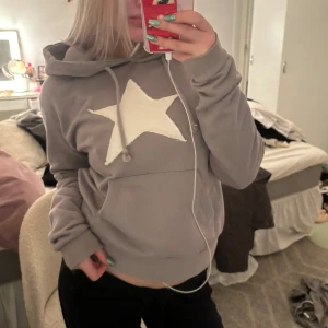 Grå hoodie med stjärna - Säljer en supermysig grå hoodie med en stor vit stjärna på framsidan. Den har en känguruficka och justerbar huva med snören. Perfekt för chilliga dagar eller när du bara vill vara bekväm. Passar till allt och är i bra skick!