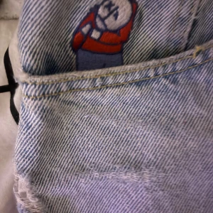 Baggy jeans från Big Boy - Säljer ett par ljusa jeans från Big Boy som inte säljs längre i storlek XS. Baggy i benen men passarxs-s i midjan. De har ett coolt broderi av en figur på fickan och en snygg slitning på ena benet. Perfekta för en avslappnad stil med en touch av personlighet. 