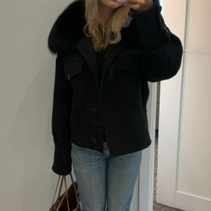 Intressekoll!! - Säljer en snygg svart jacka med pälsdetaljer på kragen. Jackan har en klassisk design med knappar framtill och två fickor på bröstet. Perfekt för höst och vinter, den är använda ett fåtal gånger och är lite nopprig men annars jätte fint skick. 
