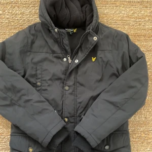 Svart jacka från Lyle & Scott Junior - Säljer en svart jacka från Lyle & Scott Junior i mycket bra skick. Jackan har en dragkedja och knappar framtill, samt en mysig huva. Perfekt för höst och vinter med sin varma och stilrena design. Passar barn i åldern 10-11 år.