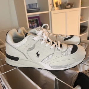 Vita sneakers från Polo Ralph Lauren - Säljer ett par snygga vita sneakers från Polo Ralph Lauren i storlek herr 45. Skorna har svarta detaljer och är tillverkade i en mix av läder och textil. Perfekta för både vardag och träning. De är i mycket bra skick och har en klassisk snörning.