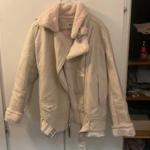 Beige jacka från Stradivarius - Säljer en superfin beige jacka från Stradivarius i storlek M. Den har en mysig fuskpälsfoder och dragkedjor på sidorna. Perfekt för höst och vinter! Jackan är i mycket bra skick och har en snygg, lite oversized passform. Perfekt för att hålla sig varm och stilren under kyliga dagar.