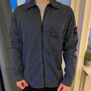 Blå overshirt från Stone Island - Säljer en snygg blå äkta overshirt från Stone Island i mycket bra skick. Den har en dragkedja framtill och en bröstficka med knapp. Perfekt för höst och vår. Passar både till vardags och lite finare tillfällen.