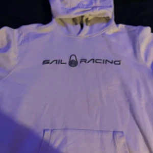 Vit hoodie från Sail Racing - Säljer en vit hoodie från Sail Racing i bra skick. Den har en klassisk design med logga på bröstet och en praktisk magficka. Perfekt för både vardag och chilliga dagar. Huvan är skön och ger extra värme när det behövs. Passar perfekt till jeans eller joggers!