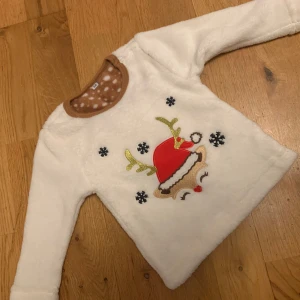 Vit jultröja med renmotiv - Supergullig vit tröja i mjukt material med ett broderat renmotiv i rött och guld. Perfekt för julen med snöflingor som detaljer. Tröjan har långa ärmar och en mysig brun krage med vita prickar på insidan. Perfekt för att hålla de små varma under vintern! 🎄