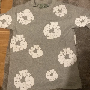 Grå t-shirt med blommönster från Denim Tears - Säljer en grå t-shirt från Denim Tears med ett coolt vitt blommönster över hela. Perfekt för en avslappnad stil. T-shirten är i bomull och har en normal passform. Passar perfekt till både jeans och shorts!