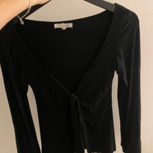 Svart långärmad topp från Design by Si - Snygg svart långärmad topp från Design by Si i storlek XS/S. Toppen har en v-ringad design med knytning framtill, vilket ger en elegant touch. Perfekt för både vardag och festliga tillfällen. 