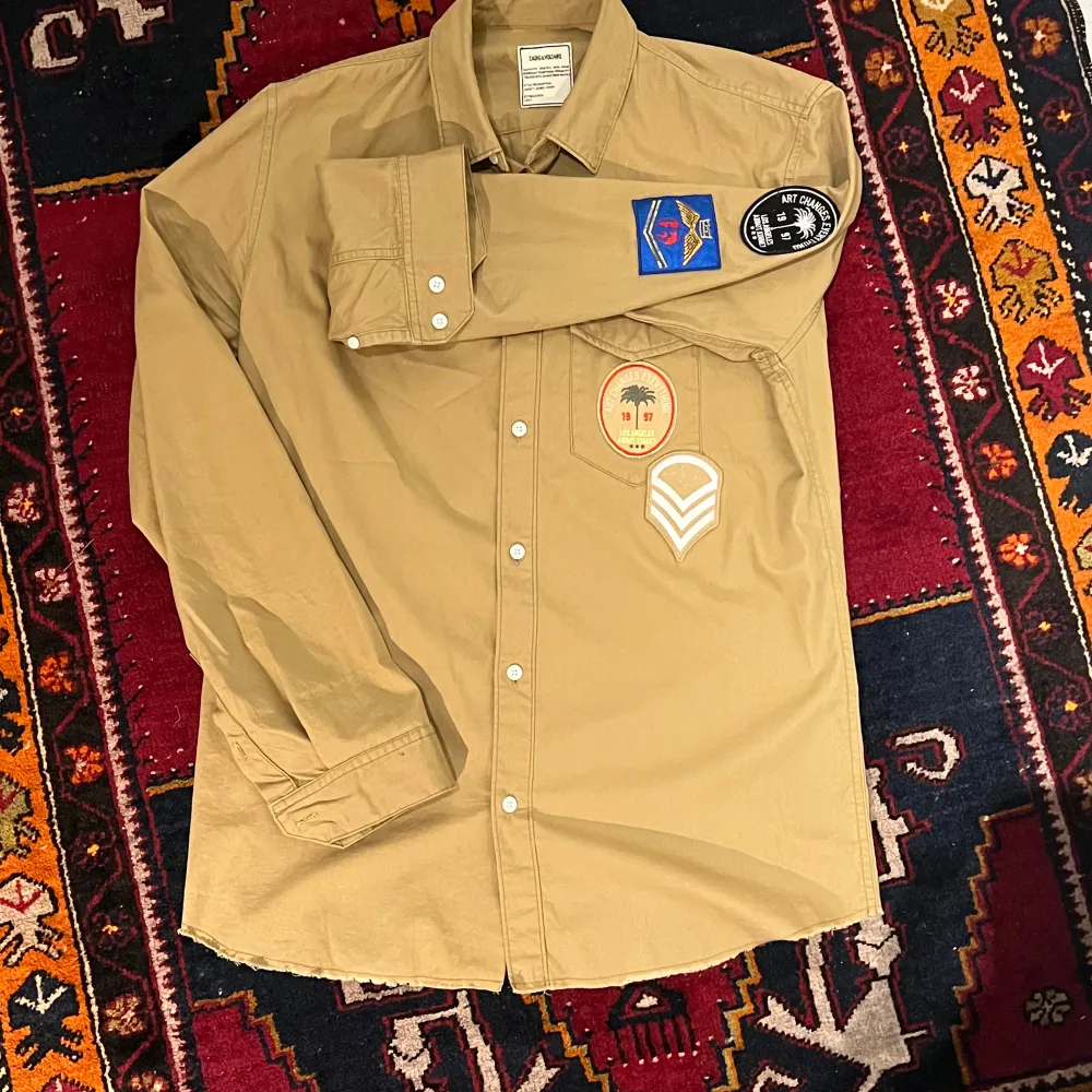 Säljer en beige långärmad skjorta från Zadig & Voltaire. Den har coola patches på bröstet och ärmen, vilket ger den en unik stil. Skjortan är i bomull och har knappar hela vägen ner. Perfekt för en avslappnad men ändå stilren look. Passar bra till både vardag och fest! Nypris cirka 1800kr. Skjortor.