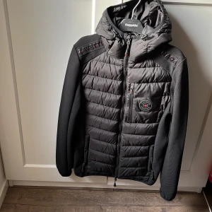 Svart cardigan från Zavetti Canada - Säljer en svart pufferjacka från Zavetti Canada i storlek XS. Jackan har en quiltad design med dragkedja och en praktisk huva. Perfekt för kyliga dagar och har en snygg logga på bröstet. Passar både till vardags och lite mer uppklädda tillfällen.