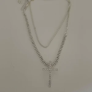 Dubbelkedjat halsband med korshänge - Snyggt dubbelkedjat halsband i silver med ett glittrigt korshänge. Perfekt för att ge en extra touch till din outfit, oavsett om det är till vardags eller fest. Kedjorna är i olika längder för en trendig lager-på-lager-look.