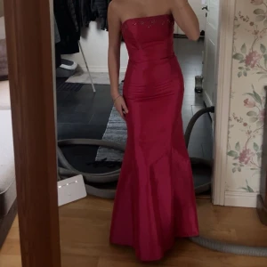 Röd axelbandslös långklänning - Säljer en elegant röd axelbandslös långklänning med glittriga detaljer upptill. Perfekt för fest eller bal! Klänningen har en tight passform som framhäver figuren och är i mycket bra skick. Materialet är glansigt och ger en lyxig känsla. Jag är 168. Priset går att diskutera 🥰