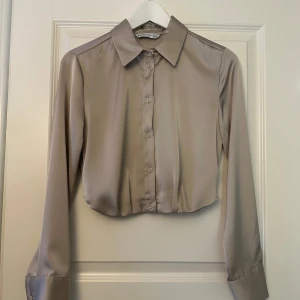 Beige croppad siden skjorta från Stradivarius - Säljer en snygg beige croppad siden skjorta från Stradivarius. Den har långa ärmar och knappar framtill. Perfekt för en stilren look, oavsett om det är till vardags eller fest. Materialet känns mjukt och lyxigt, vilket gör den bekväm att bära hela dagen.