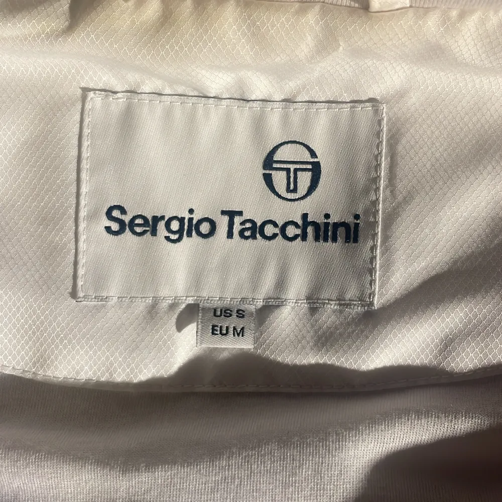 Säljer en snygg vit och blå vindjacka från Sergio Tacchini. Jackan har ett coolt rutmönster på den vita delen och en dragkedja framtill. Den har också en huva och elastiska ärmslut. Perfekt för vår och höst! Pris går alltid att förhandlas! Andvänd en handfull gånger Max!. Jackor.