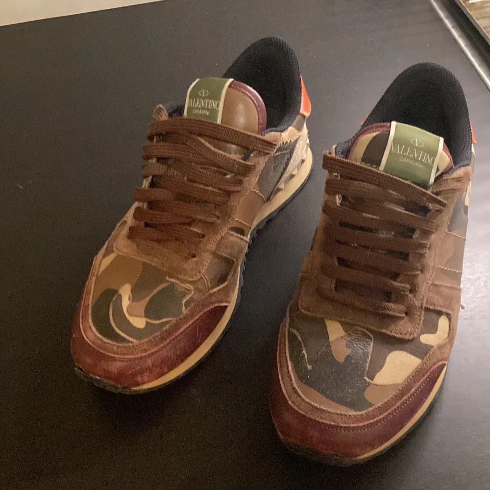 Säljer ett par Valentino rockrunners i camouflage-mönster. Skorna har bruna och beige toner med en touch av rött och orange på hälen. De är i bra skick och har snörning framtill. Storlek 40. Skor.