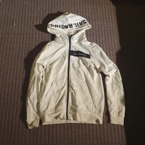 Vit huvtröja från Sail Racing (äkta) - Säljer en snygg vit huvtröja från Sail Racing i nytt skick. Den har en dragkedja framtill och en stor logga på huvan. Perfekt för vår och höst, med långa ärmar och en skön passform. Passar både till vardags och lite sportigare tillfällen.