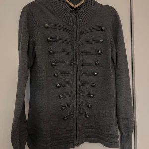Grå kofta från Promiss - Säljer en snygg grå kofta från Promiss i storlek L. Den har en cool militärinspirerad design med dubbla rader av knappar framtill och en dragkedja. Perfekt för höst och vinter, och den är i mycket bra skick. Passar både till vardags och lite finare tillfällen.