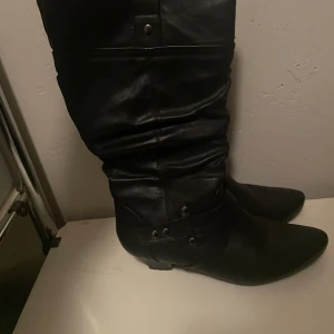 Svarta stövlar i skinn - Säljer ett par svarta stövlar i skinn med en stilren design. De har en spetsig tå och dekorativa detaljer med nitar. Perfekta för höst och vinter, dessa stövlar är både snygga och praktiska.