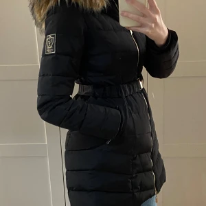 Chiara Forthi jacka - Svart jacka med avtagbar pälskrage. Storlek 38. Jackan har en dragkedja framtill och en bälte i midjan. Jackan är i helt ny skick, aldrig använd.