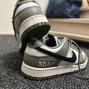 Nike Dunk Low NBA Edition - Säljer ett par Nike Dunk Low i NBA Edition. Skorna är i grönt och vitt med svarta detaljer och har en snygg NBA-logga på hälen. De är i mycket bra skick och har klassisk snörning. Perfekta för både vardag och sportiga tillfällen! Väldigt snålt använt (enstaka gånger). Köparen står för frakt. 