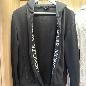 Svart hoodie från Moncler - Säljer en svart hoodie från Moncler i mycket bra skick. Den har en dragkedja framtill och långa ärmar. Moncler-loggan syns på insidan av luvan. Inköpt jul 2020. Nypris 13000. Kvitto finns.