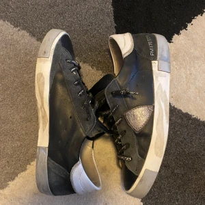 philippe modell skor - Säljer ett par svarta sneakers med vita detaljer och coola snören med text. Skorna har en sliten look men är fortfarande i bra skick. Perfekta för en avslappnad stil och passar till de flesta outfits. De har en bekväm passform och är gjorda i skinn. Strl 40 men passar 40,5 oxå|| Pris kan diskuteras vid snabb affär🤝
