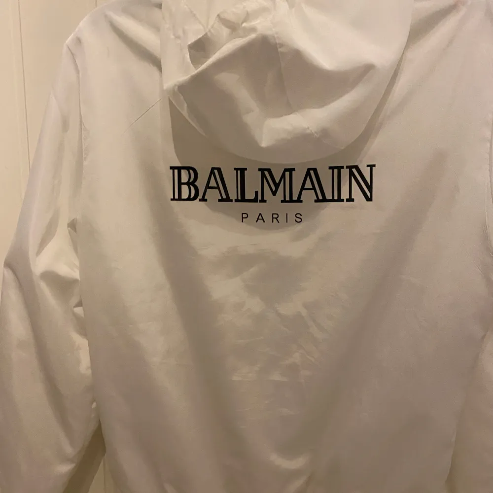 Säljer en stilren vit vindjacka från Balmain. Jackan har en dragkedja framtill och en huva för extra skydd mot väder. Balmain-loggan är tryckt både fram och bak. Perfekt för vår och höst! Storlek S . Jackor.