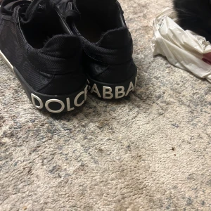 Svarta sneakers från Dolce & Gabbana - Säljer ett par svarta sneakers från Dolce & Gabbana i bra skick. Skorna har en vit sula och tydlig logga baktill. De är tillverkade i syntetmaterial med snörning framtill. Perfekta för en stilren vardagslook!