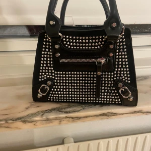 Svart handväska med nitar - Säljer en liten svart handväska. Den liknar Balenciagas väskor. Finns ett längre band som man kan fästa på väskan om man vill. Nyskick använd 3 gånger.