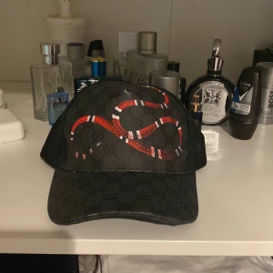 Svart keps med ormmönster från Gucci - Säljer en svart keps från Gucci med ett coolt ormmönster i rött och vitt. Kepsen har justerbart band bak med blå och röda ränder. Perfekt för att ge din outfit en lyxig touch. Passar både till vardags och fest!