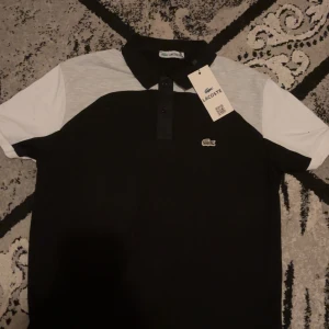 Svart och vit pikétröja från Lacoste - Säljer en snygg pikétröja från Lacoste i svart och vitt. Tröjan har en klassisk krage med knappar och den ikoniska krokodilloggan på bröstet. Perfekt för både vardag och fest! 🐊